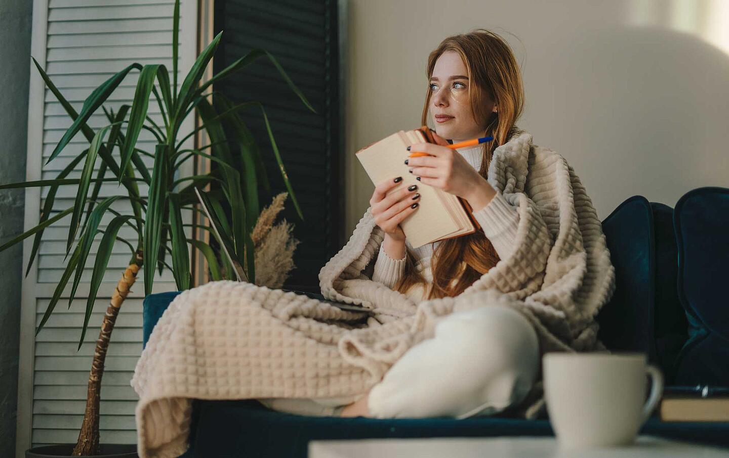 Resilienz: Junge Frau, eingewickelt in eine Decke, sitzt auf dem Sofa