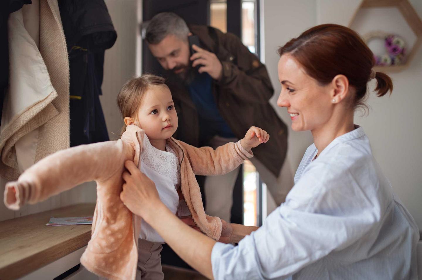 Mutter zieht kleiner Tochter die Jacke an, im Hintergrund telefoniert der Vater
