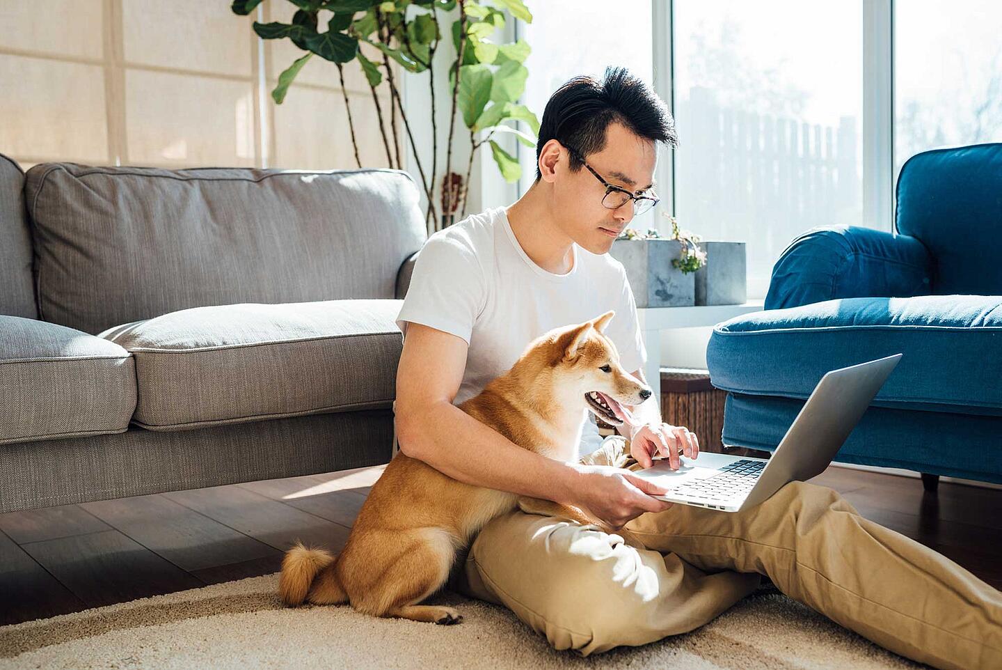 Work-Life-Blending – Mann sitzt im Wohnzimmer auf dem Fußboden und arbeitet mit Hund im Arm am Laptop