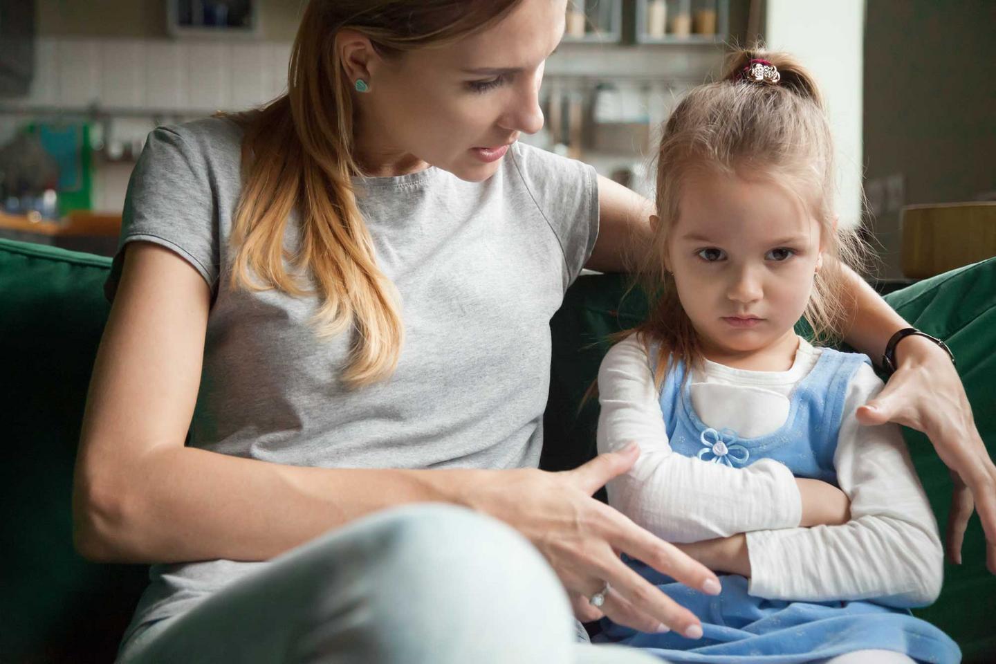 Mutter und ihre kleine Tochter sitzen auf dem Sofa, die Mutter hat einen Arm um das Mädchen mit trotzigem Gesichtsausdruck gelegt und sprcht mit ihr